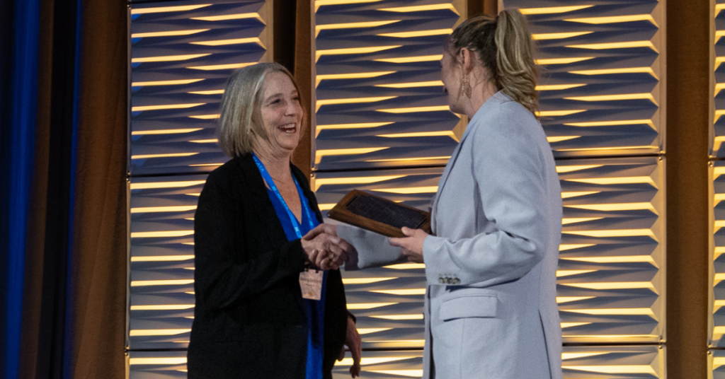 Image of woman in a grey blazer handing off an award to a woman in a black sweater