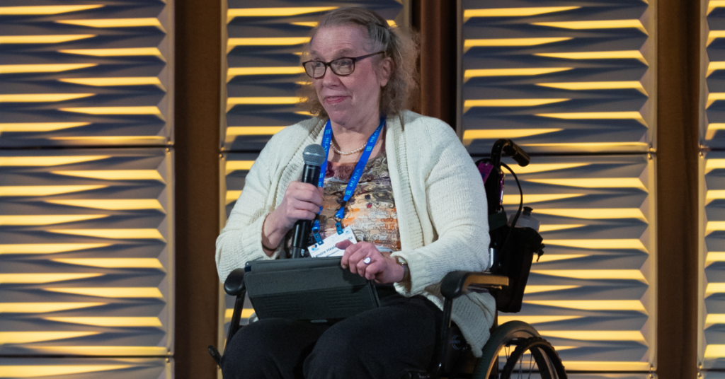 Image of a smiling woman sitting in a wheelchair and speaking into a microphone
