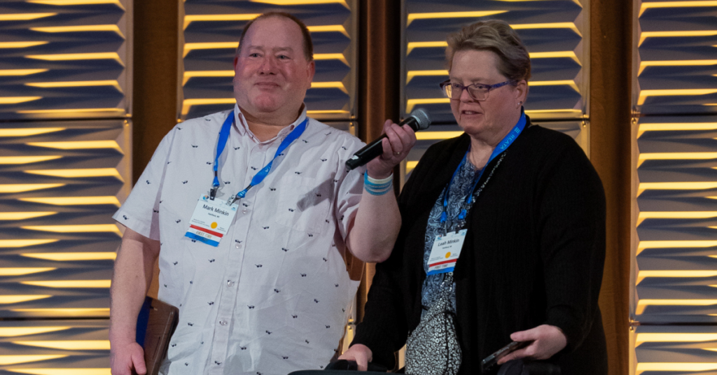 Image of a man in a blue collar shirt holding an award and a woman standing next to him in a black sweater speaking into a microphone