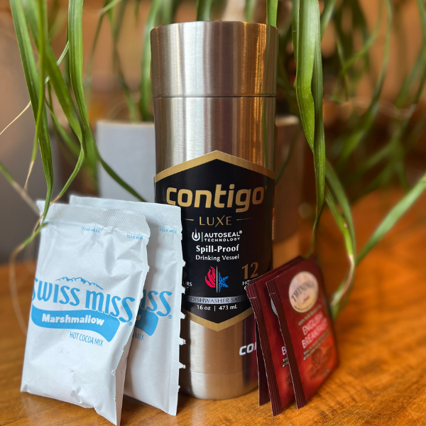 A Silver Contigo Luxe Autoseal Travel Mug on a wooden table with Swiss Miss hot chocolate packets and tea bags leaning against it and a plant behind it.