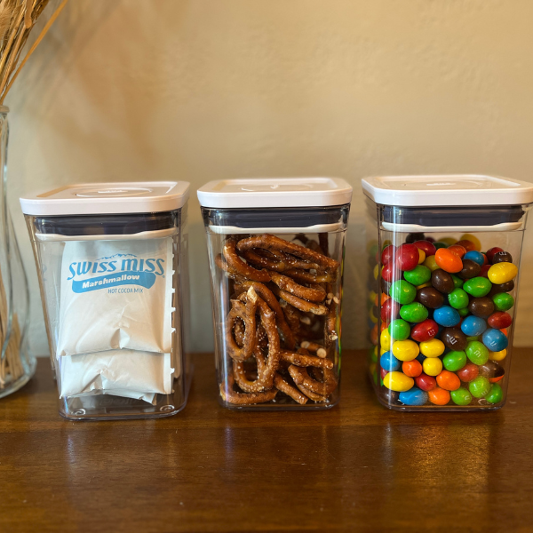 Three clear OXO POP containers are sitting on a wooden table. One is filled with hot chocolate packets, another with pretzels, and the third with colorful peanut M&Ms.